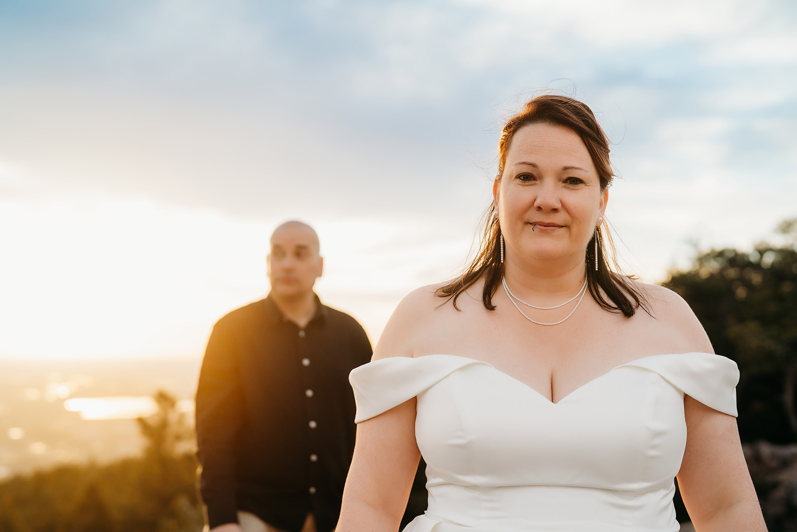 Bride walks confidently toward the camera, sunlight pouring in behind her, with her groom watching proudly from the background.