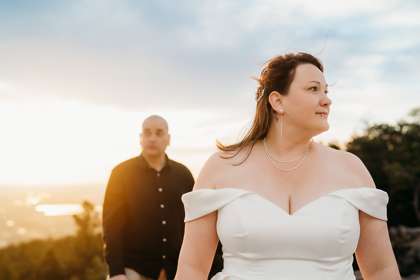 Bride gazes confidently into the distance as her groom walks behind her—sunlight glowing between them.