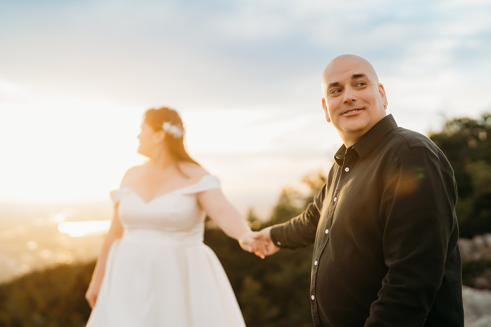 Groom smiles and looks back as he leads his bride through the mountaintop terrain, hand in hand at golden hour.