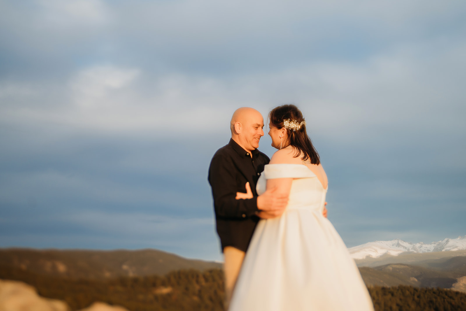 The couple locks eyes as golden hour glows around them, smiling in the breeze with snowy peaks in the distance.