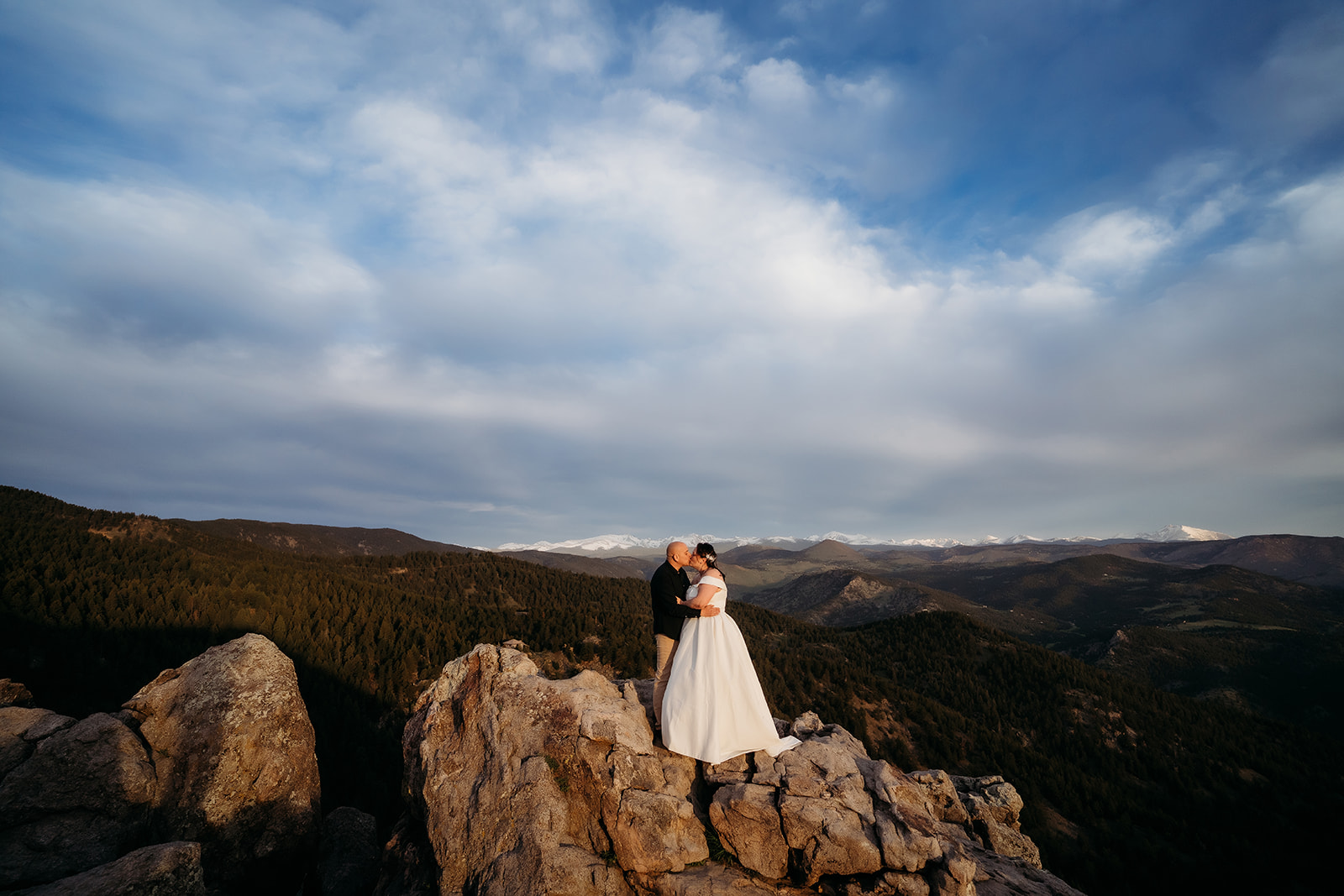 The couple embraces on a rocky overlook, surrounded by sweeping mountain views and golden hour skies.