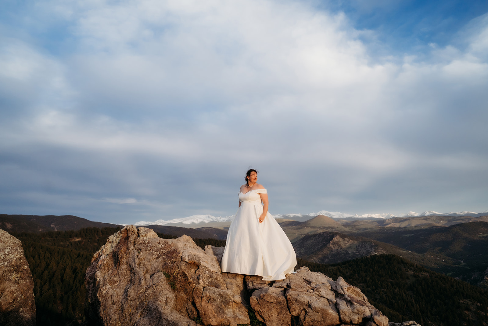 Bride stands confidently atop a rugged cliffside, dress flowing in the wind, captured by an elopement photographer Colorado.