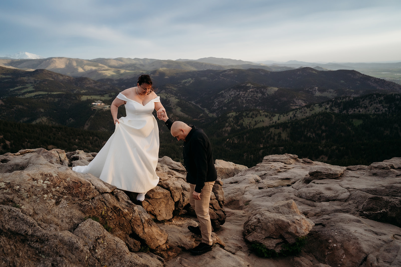 Groom helps his bride navigate rocky terrain in her gown, surrounded by Colorado’s stunning mountain views and documented by an elopement photographer Colorado.