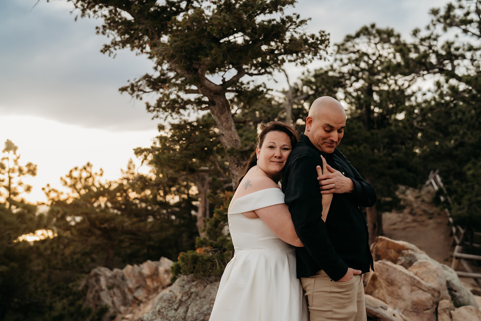 Bride wraps her arms around the groom from behind, both smiling softly against the backdrop of pine trees and golden-hour light.