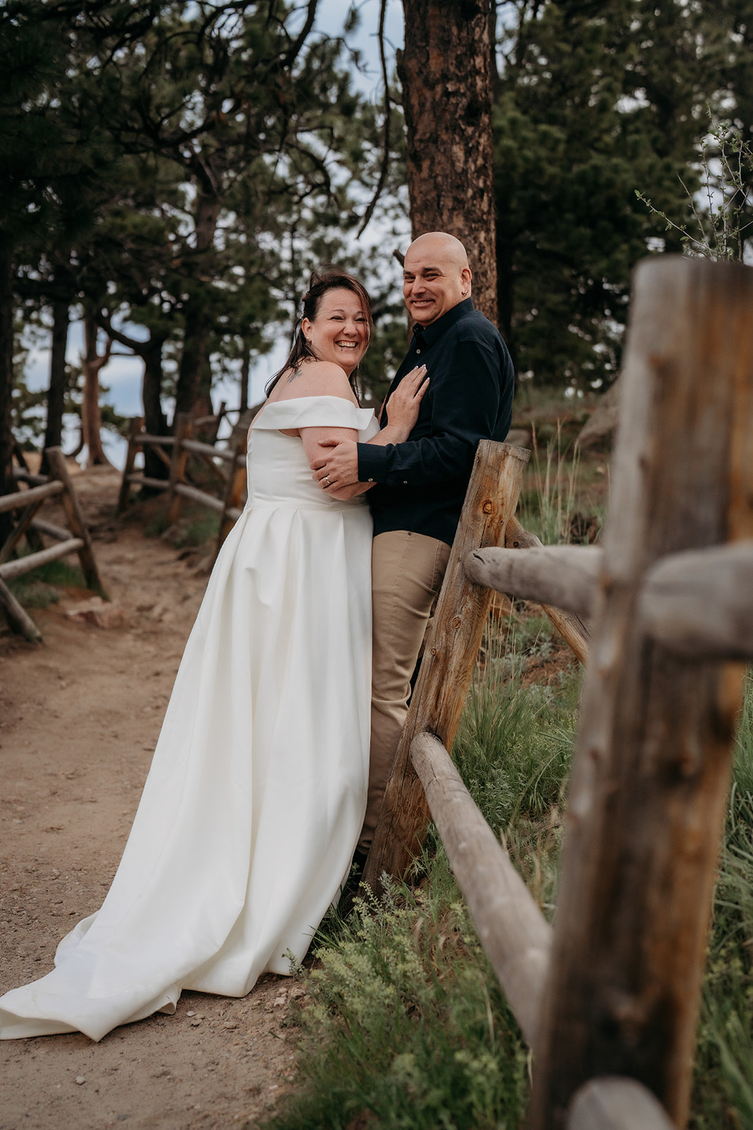 Bride and groom share a playful moment leaning against a wooden fence, laughing together in the forest.