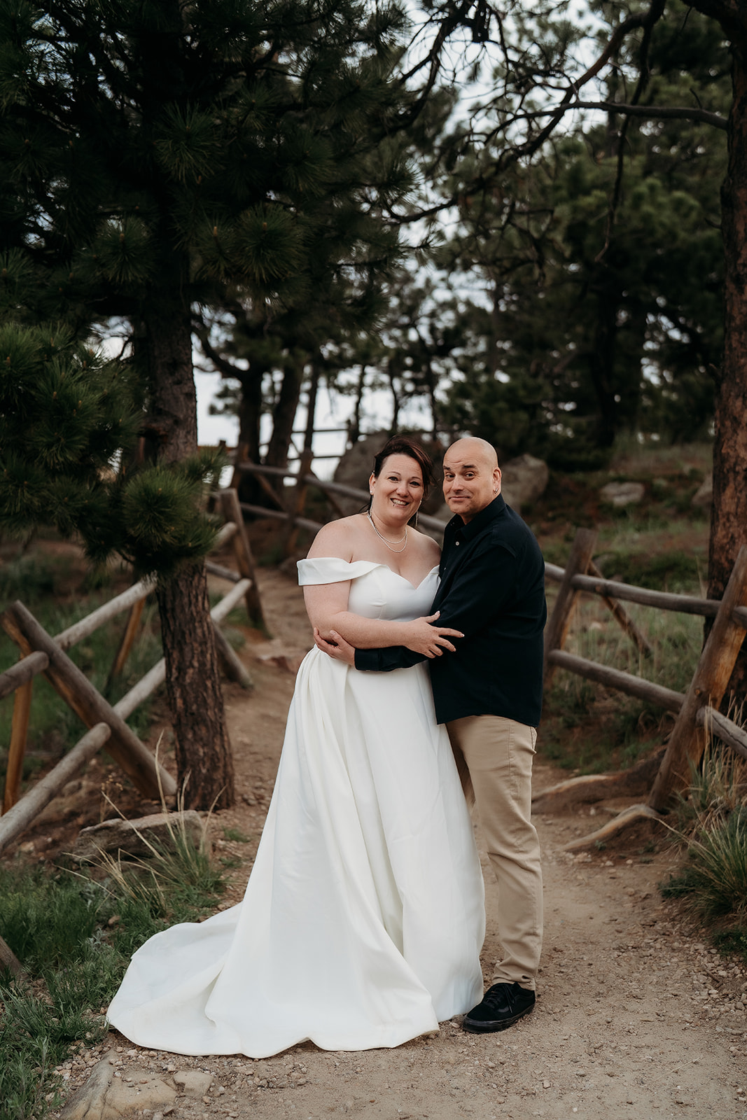 Arm in arm, the newlyweds smile at the camera, framed by tall pines and rustic wooden railings.