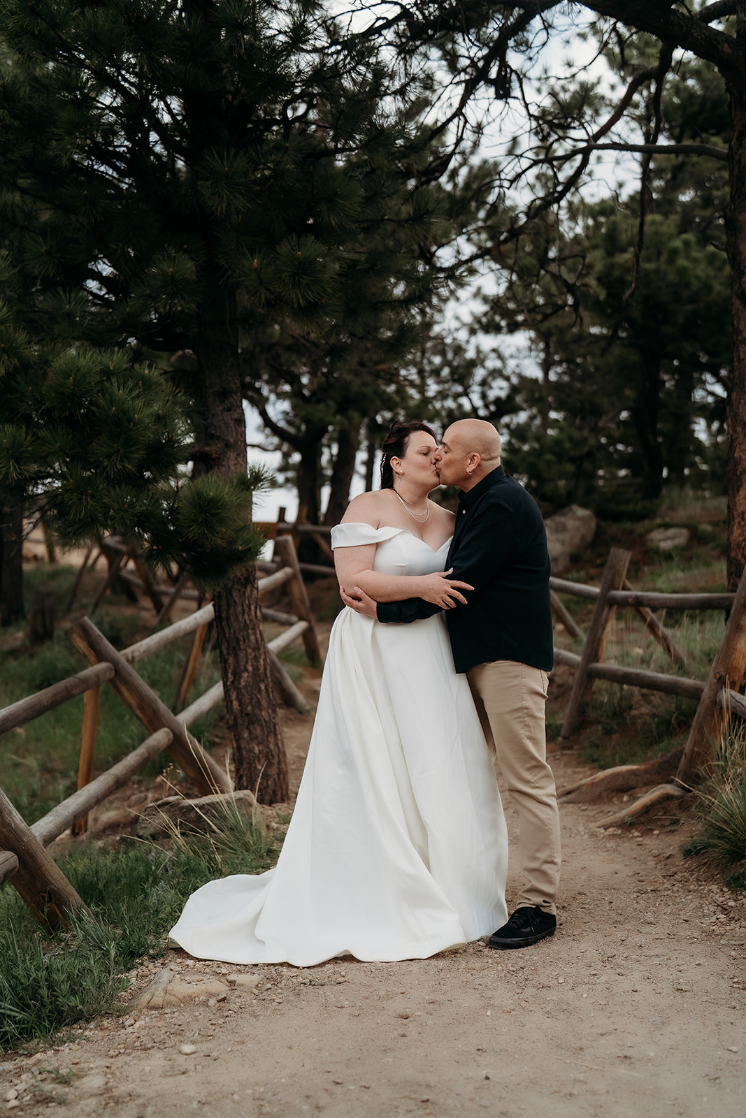 Couple kisses under the trees along a quiet trail, surrounded by rustic fencing and golden light.