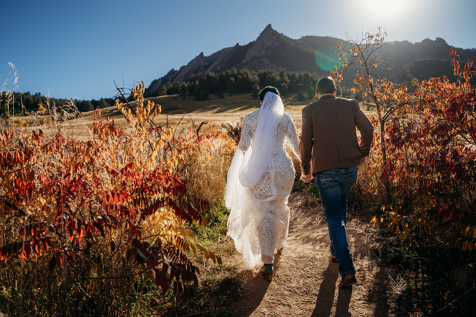 Couple walking hand-in-hand down a trail surrounded by golden and red fall foliage, with the Flatirons glowing in the background.