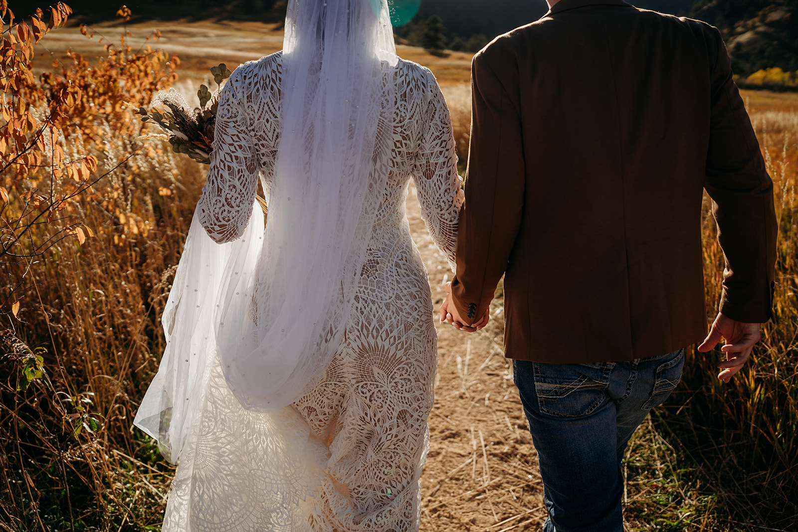 Close-up of the couple walking hand-in-hand down a golden trail, the bride’s lace gown and veil glowing in the autumn sun.