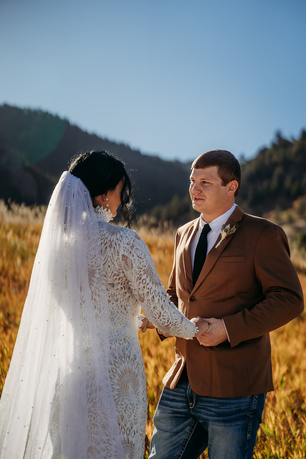 Bride and groom hold hands in a sun-drenched field, exchanging a sweet look surrounded by fall mountain colors.