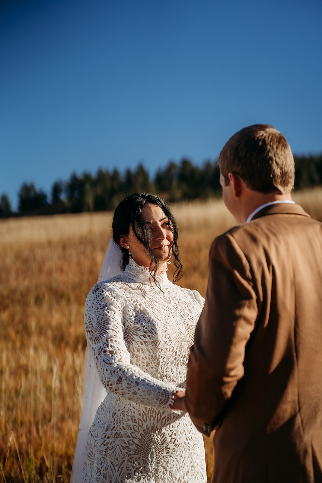 Bride gazing at her partner with emotion during their vows, standing in a quiet mountain meadow with fall grass around them.