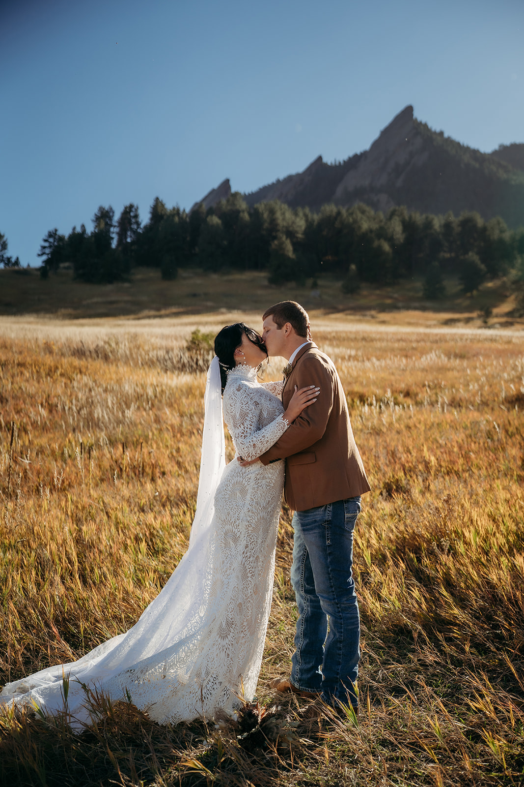 A sweet kiss shared between the newlyweds, standing in a sunlit Colorado field with dramatic mountain views behind them.