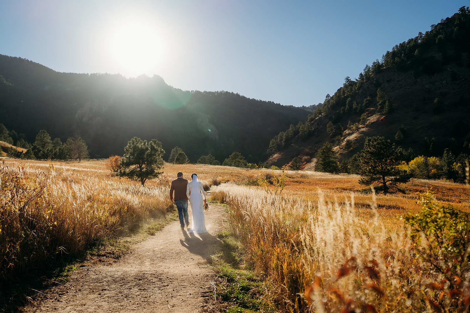 Bride and groom walk hand-in-hand along a sunlit trail surrounded by golden grass and mountain views during their Boulder elopement.