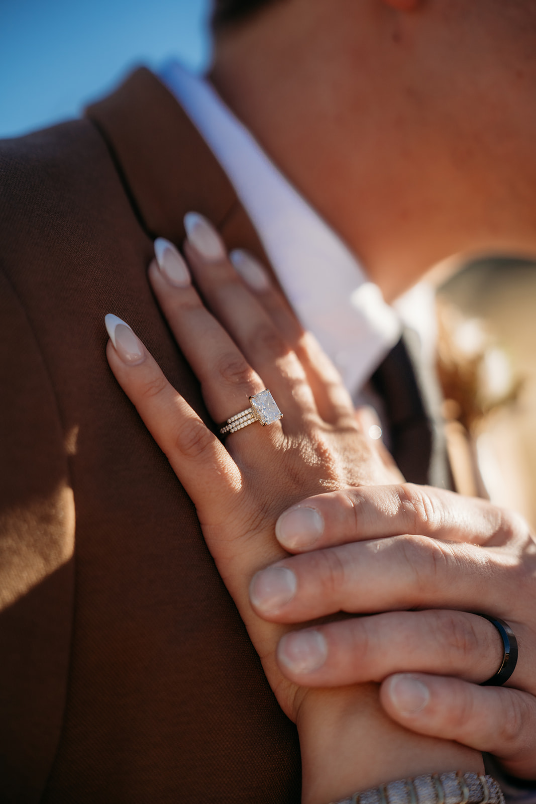 Close-up of the bride’s hand resting on the groom’s chest, her engagement and wedding rings sparkling in the sun.