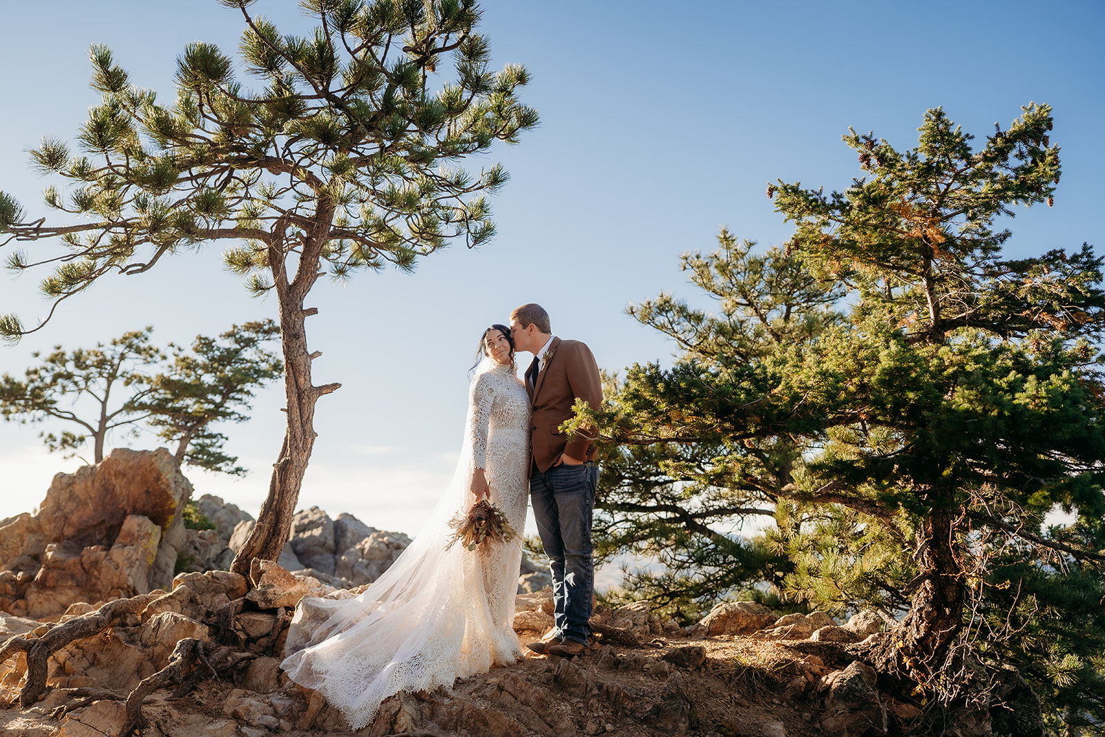 Joyful portrait of the couple embracing, framed by evergreens and soft morning light on a clear Colorado day.