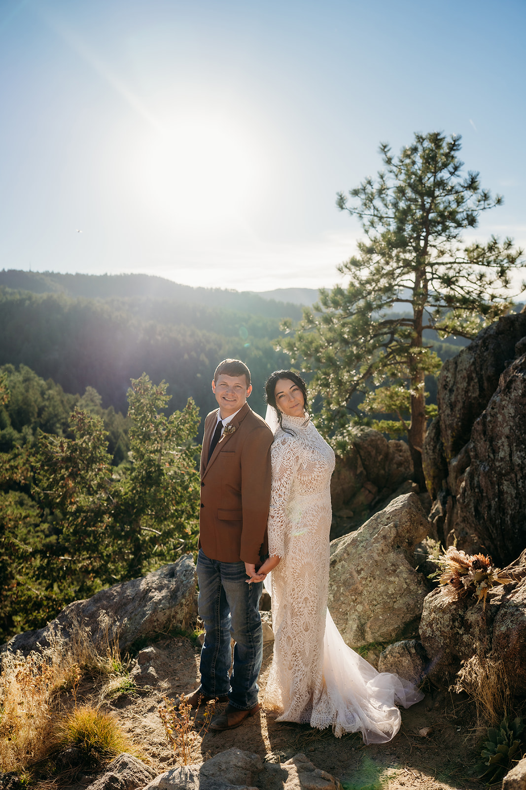 Couple holding hands and smiling in the sun, framed by forested mountains during their joyful Boulder elopement.