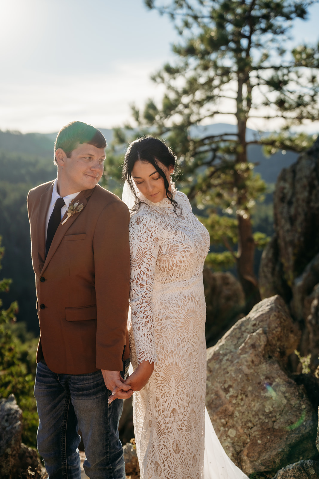 Couple sharing a quiet moment on a rocky trail, holding hands with smiles as pine trees and mountain views surround them.