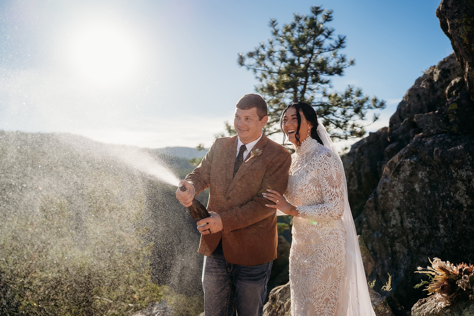 Couple pops champagne on a cliffside with sunlight sparkling around them—an untraditional, joyful start to their Boulder elopement celebration.