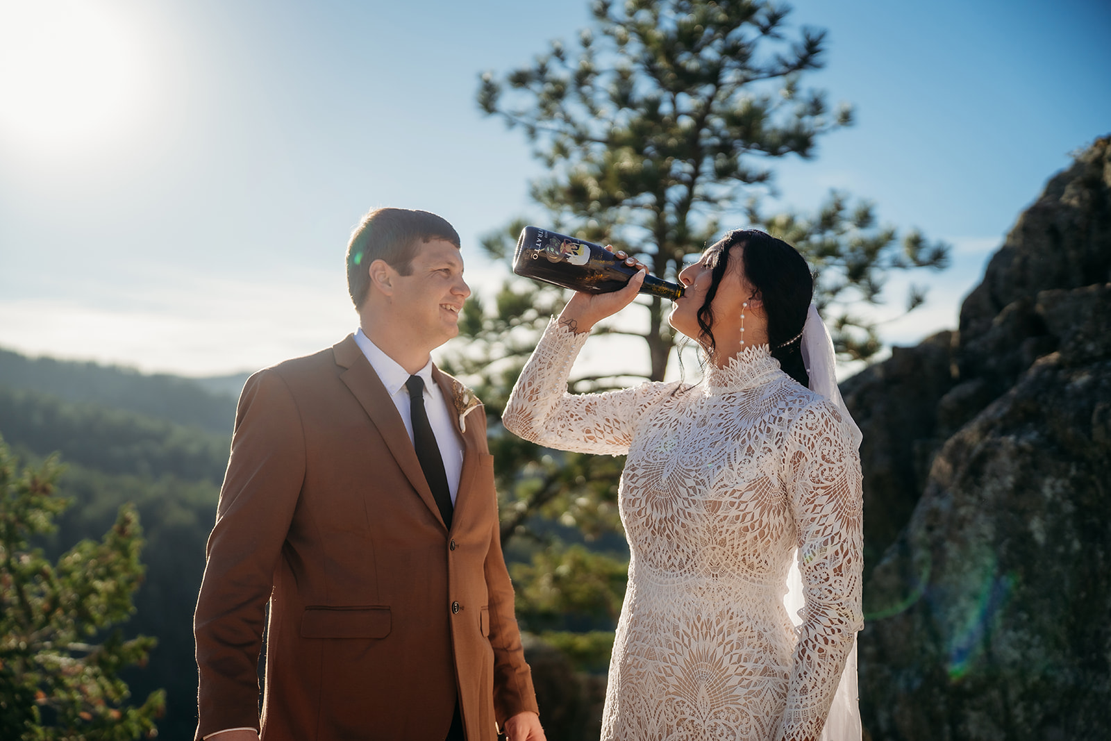 Bride takes a sip straight from the bottle, laughing with her new husband on a rocky overlook during their carefree Boulder elopement.