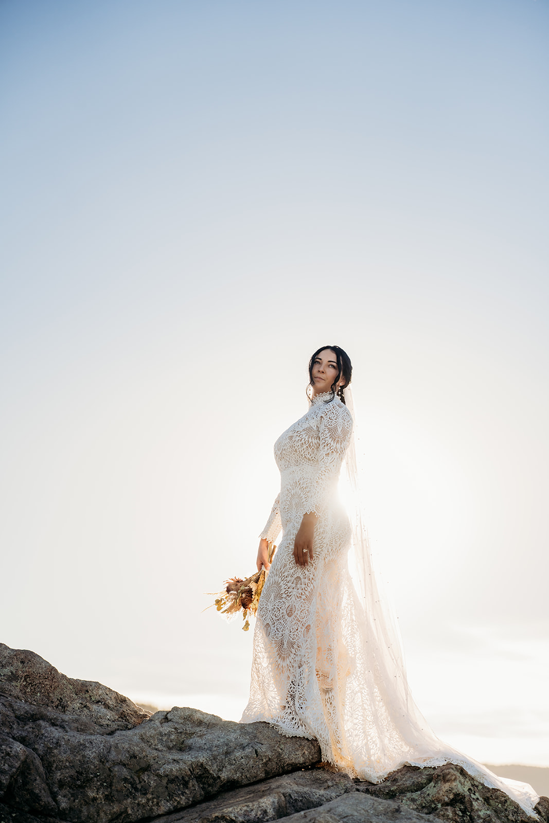 Bride standing solo on a boulder, her lace dress glowing in the golden backlight of sunset.