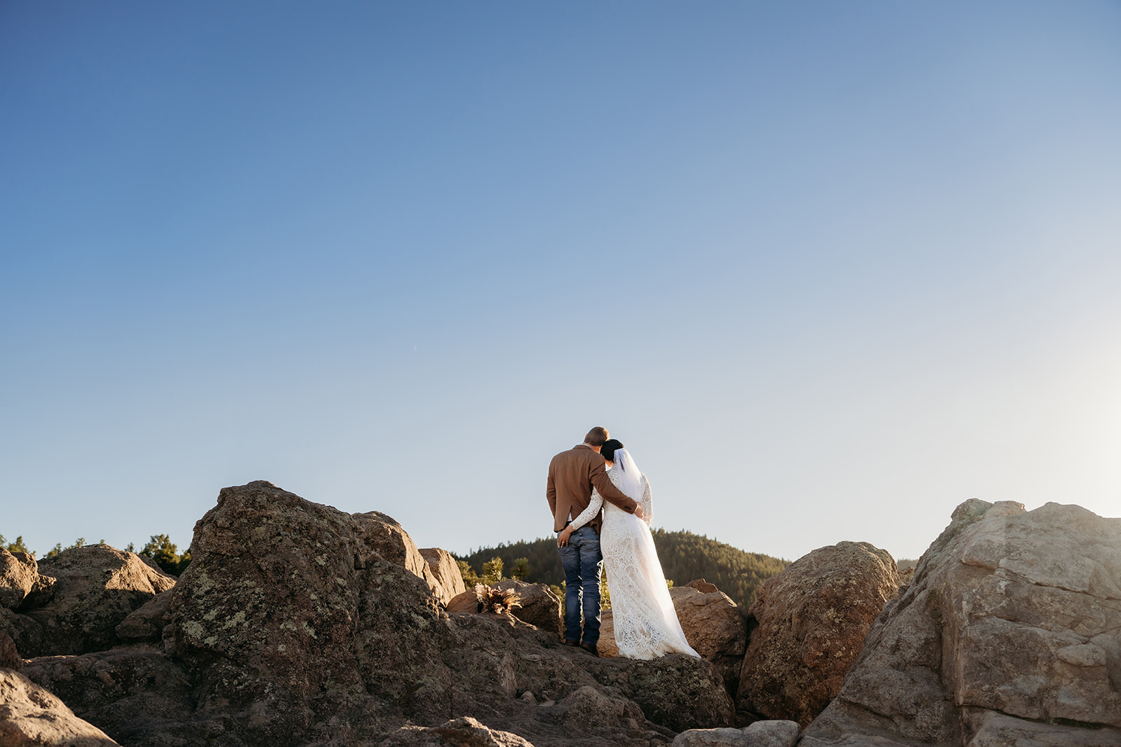Couple stands arm in arm atop a rugged rock formation, looking out over the Colorado foothills as the sun begins to set.
