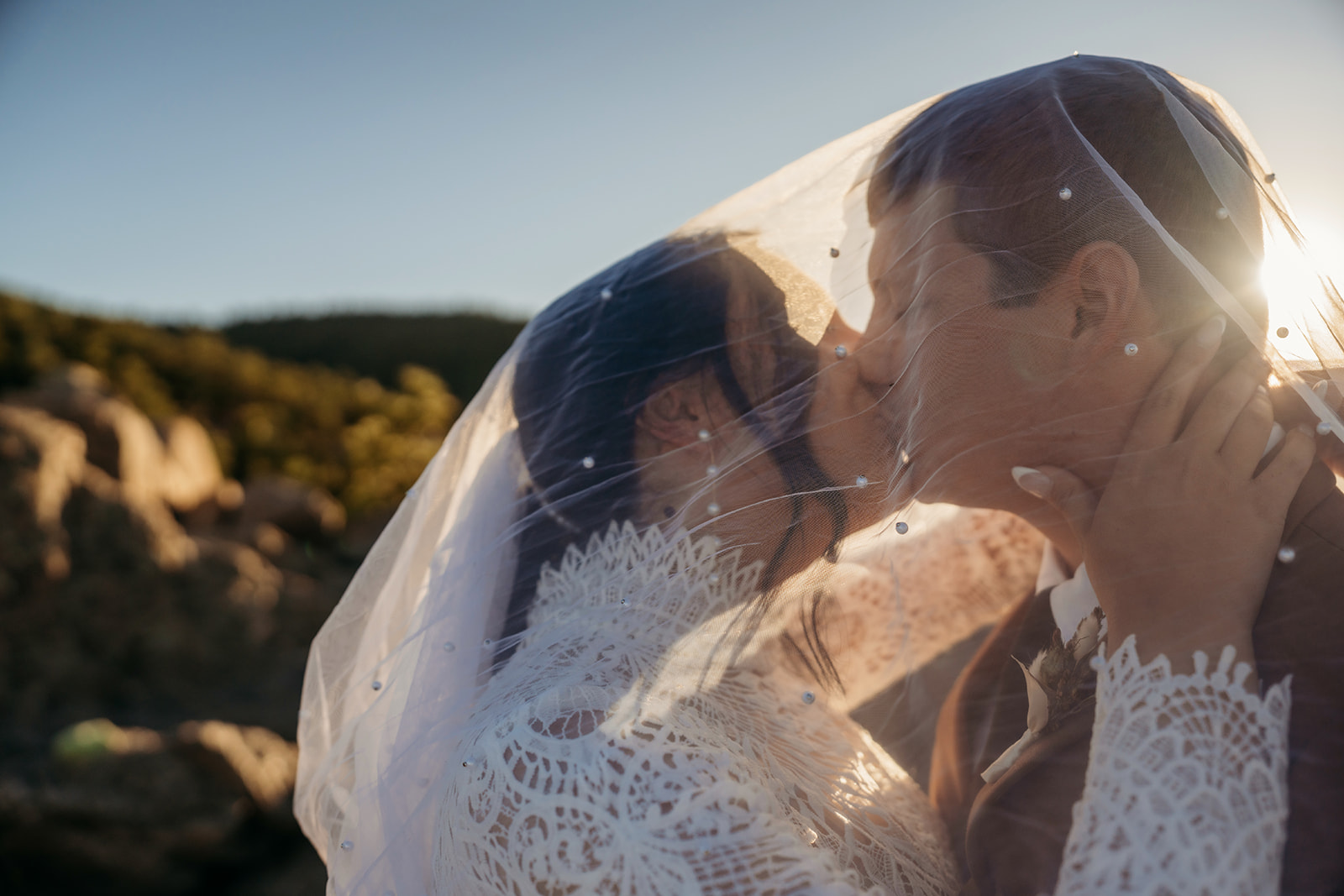 A tender kiss under the bride’s beaded veil, captured in soft golden light with mountain peaks blurred in the background.