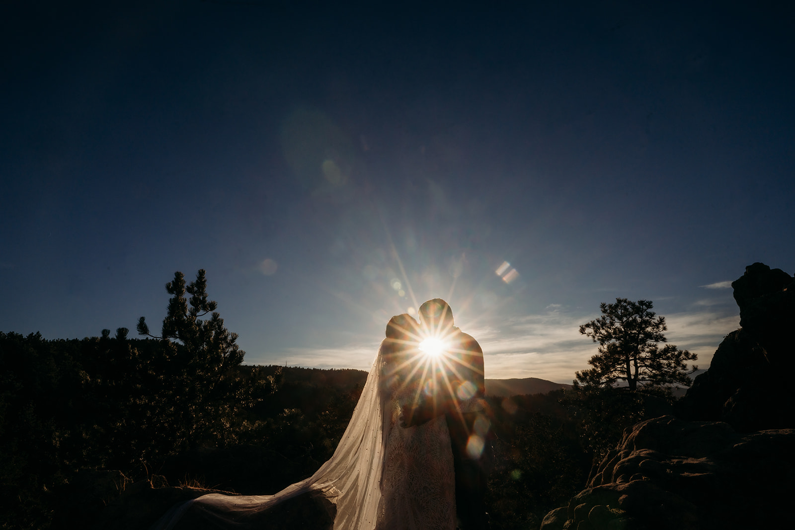 Silhouetted kiss at sunset as the sun bursts through the couple’s embrace on a rocky overlook in the Colorado mountains.