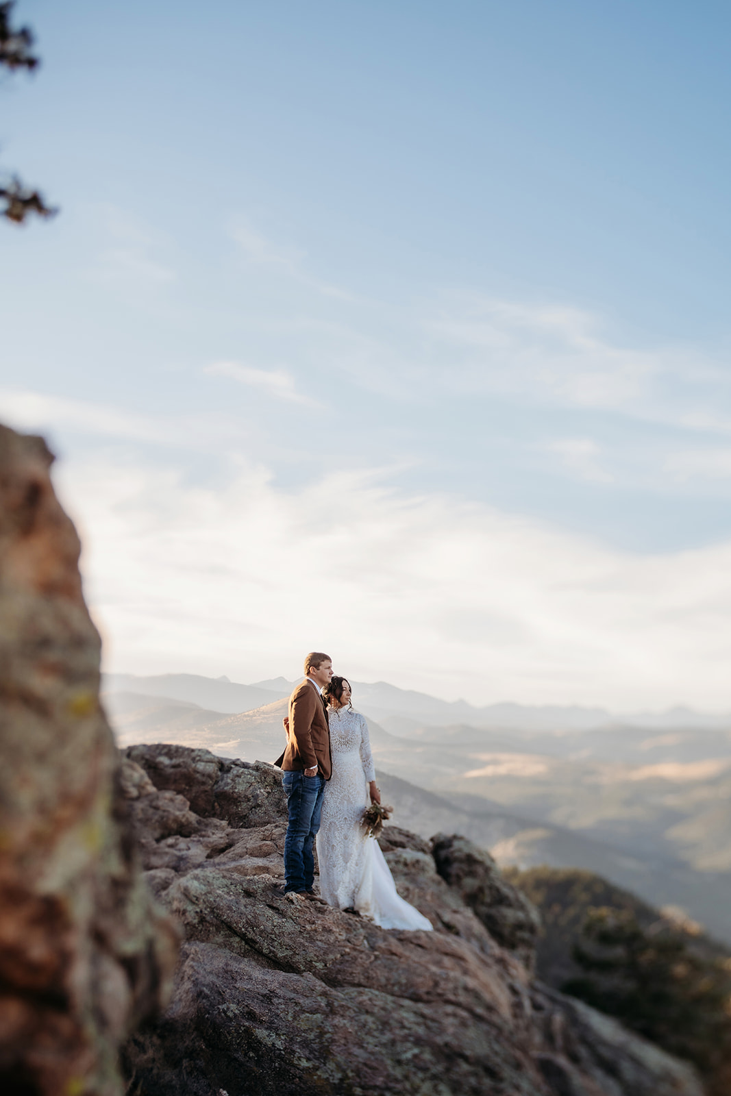 Newlyweds standing on a rocky cliff, soaking in the vast mountain views during their adventurous Boulder elopement.