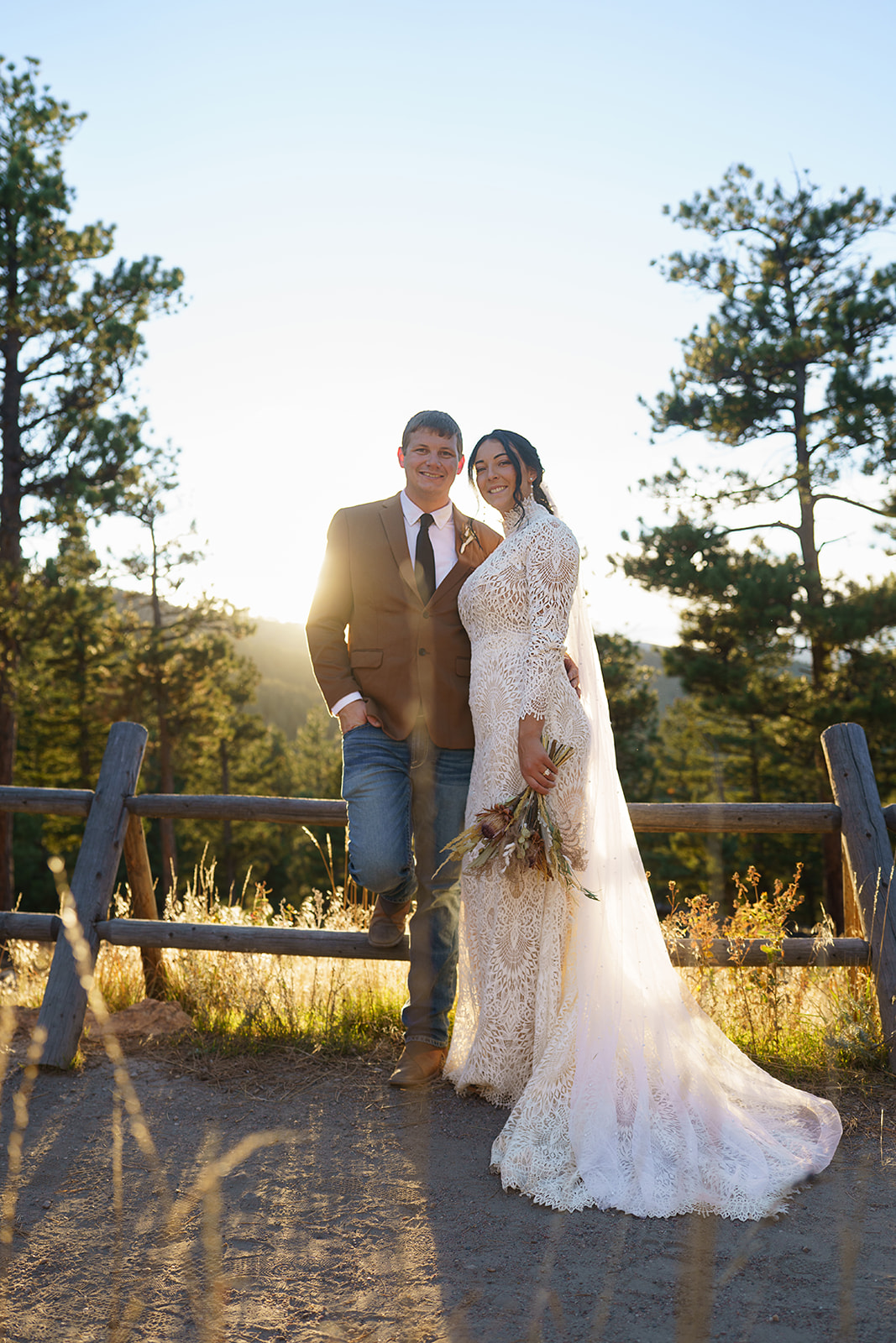 Bride and groom hold hands in a sun-drenched field, exchanging a sweet look surrounded by fall mountain colors.