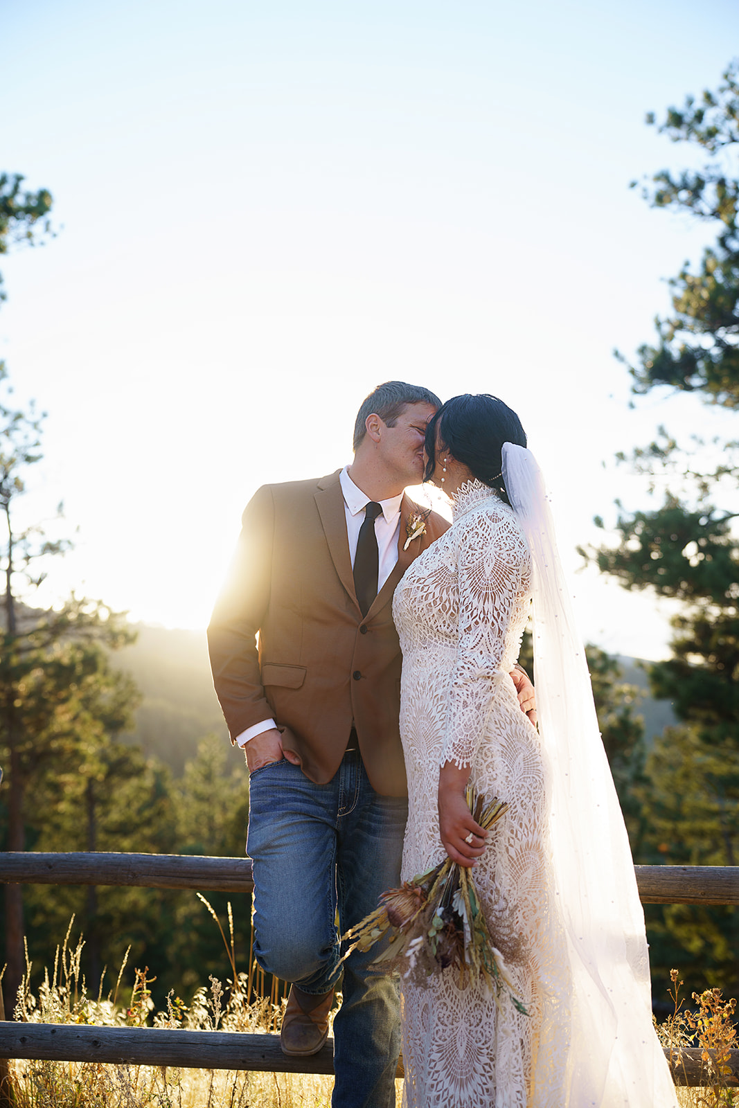 The couple shares a kiss near a wooden fence at golden hour, the bride holding a wildflower bouquet in her hand.