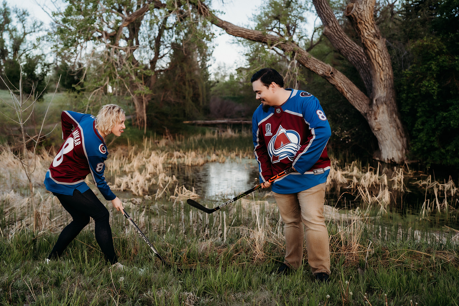 Playful moment between a couple in Colorado Avalanche jerseys, pretending to play hockey in a grassy field.