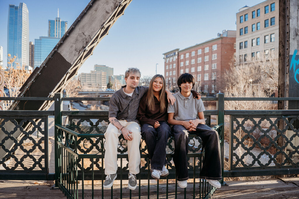 Three siblings sitting side by side on a metal railing on a Denver bridge, smiling toward the camera with the city skyline in the background during a lifestyle family photo session.
