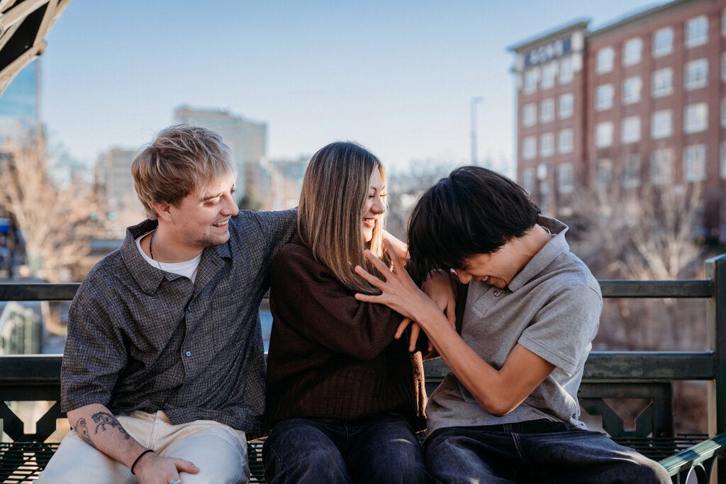 Three siblings sitting closely together on a bench on a Denver bridge, laughing and playfully interacting during a candid family session with Denver photographers.