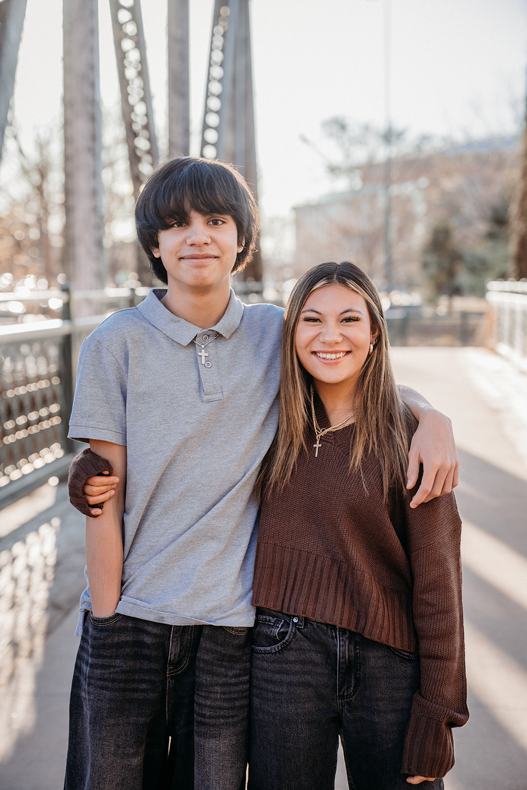 A teenage boy and young woman standing arm in arm on a Denver bridge, smiling naturally during a lifestyle family photo session.