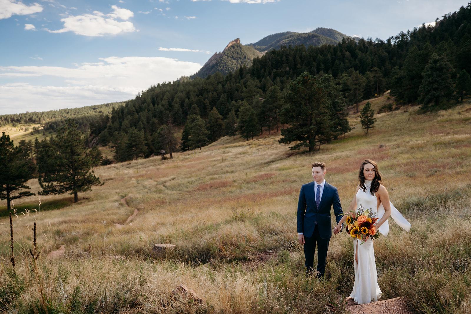 A couple standing together in a grassy mountain meadow with evergreen trees and rocky peaks in the background during Colorado weddings.
