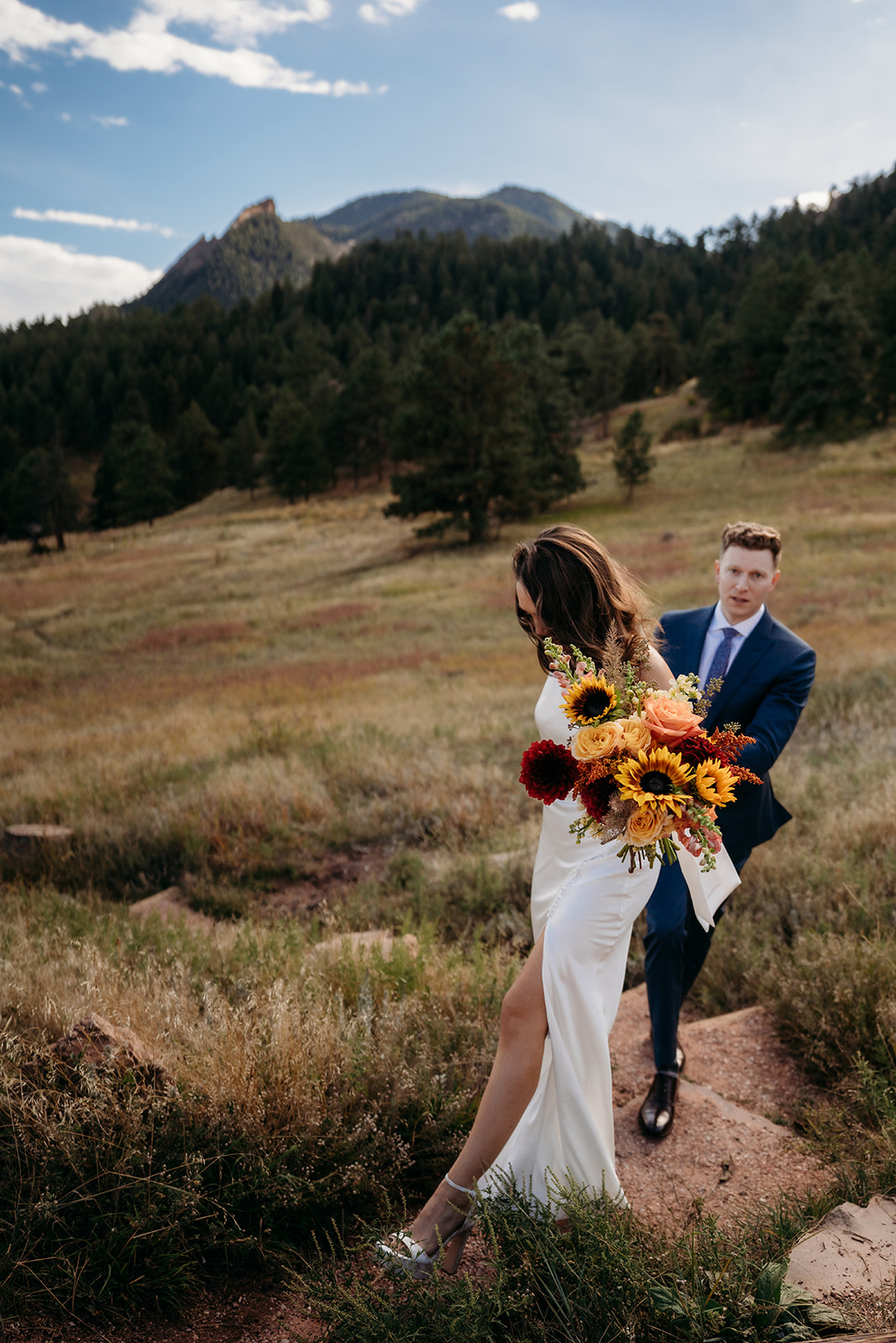 A bride walking ahead on a mountain trail while holding a bouquet, with her partner following behind during a scenic Colorado wedding day.