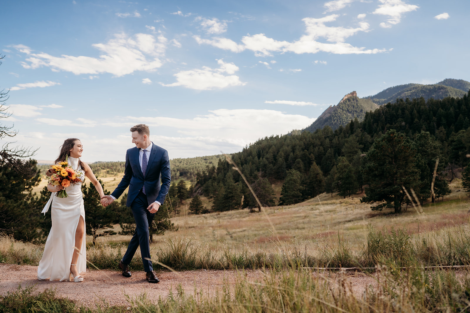 A bride and groom walking hand in hand along a trail in the Colorado foothills, smiling together during their wedding day.