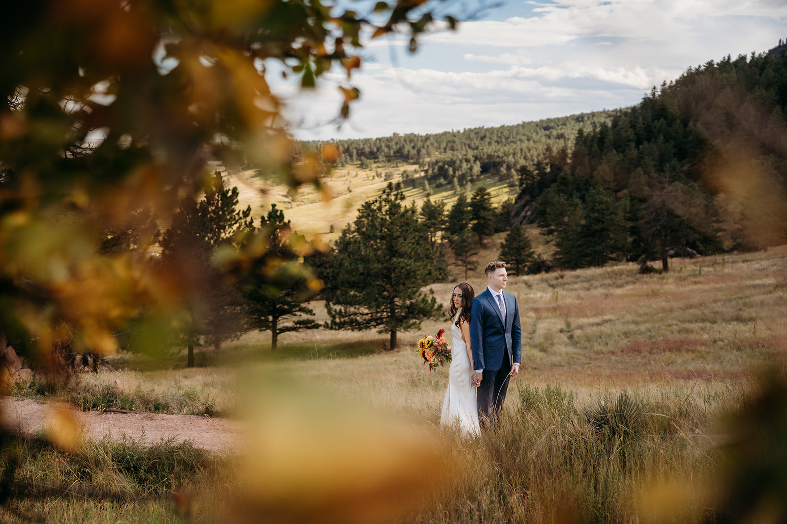 A bride and groom walking together through a grassy meadow in the Colorado foothills, framed by fall leaves during their wedding day.