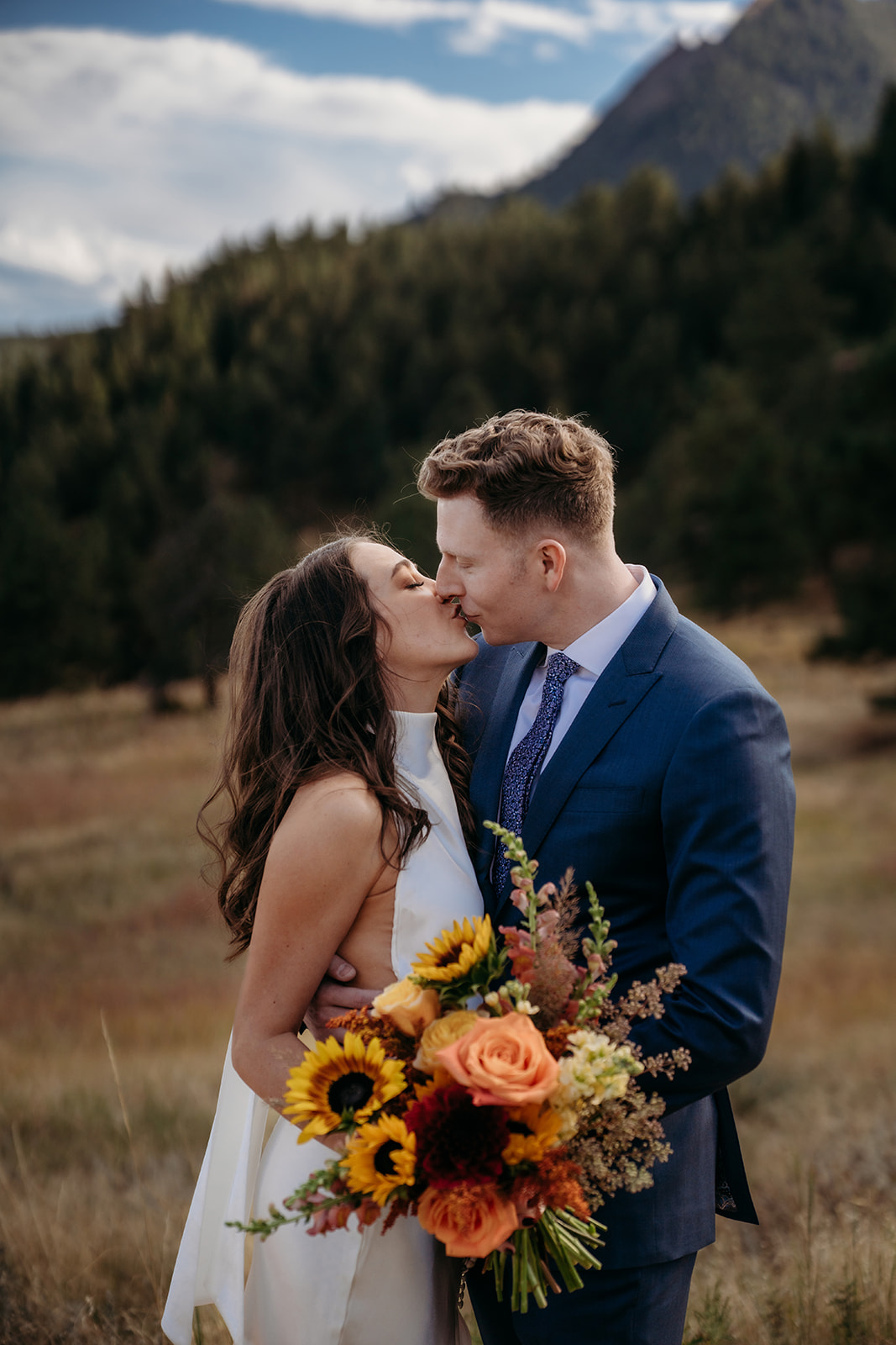 A bride and groom kissing while holding a sunflower-filled bouquet, surrounded by Colorado mountain scenery during their wedding.