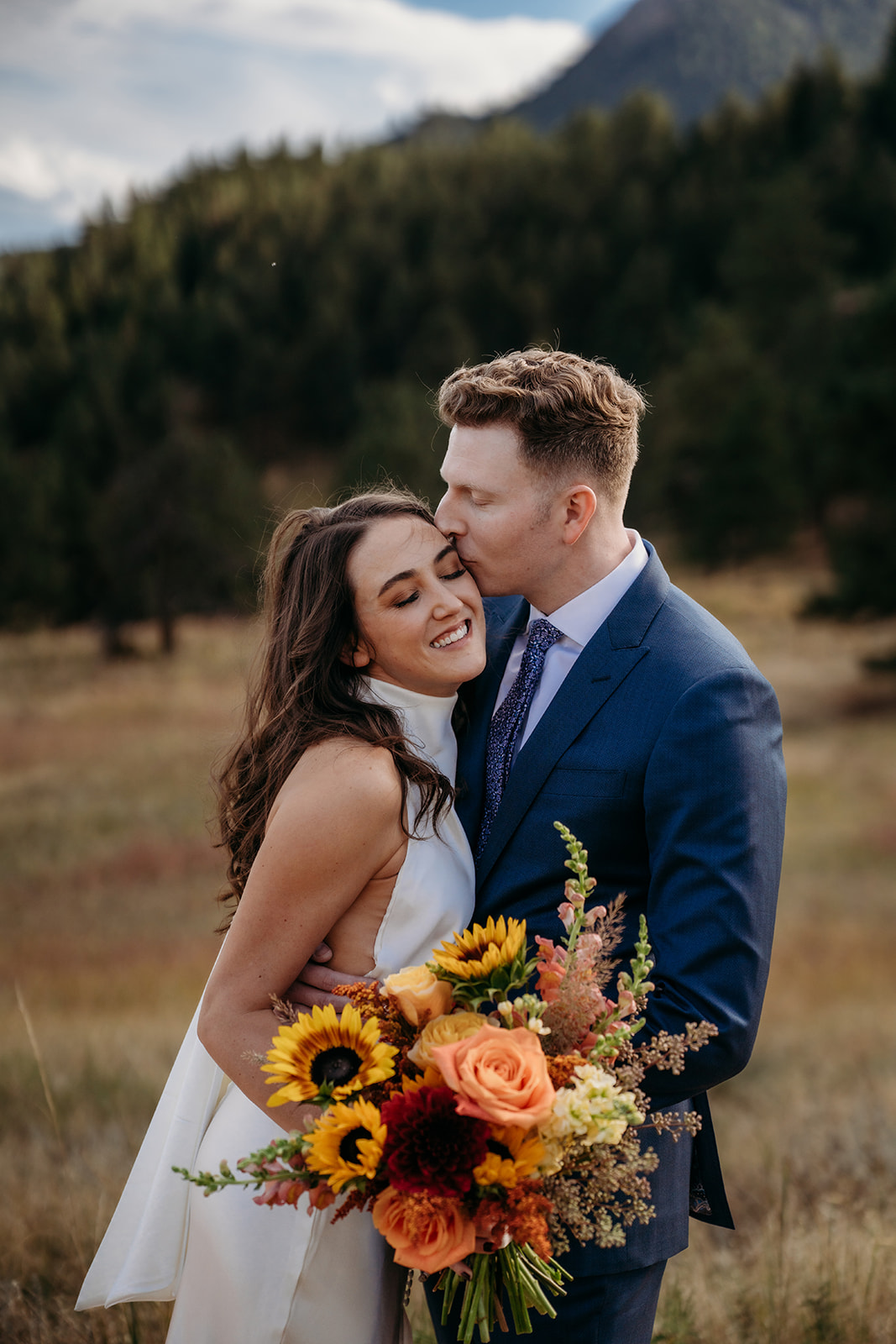 A groom kissing his bride on the cheek as she smiles while holding a vibrant fall bouquet during their Colorado wedding day.
