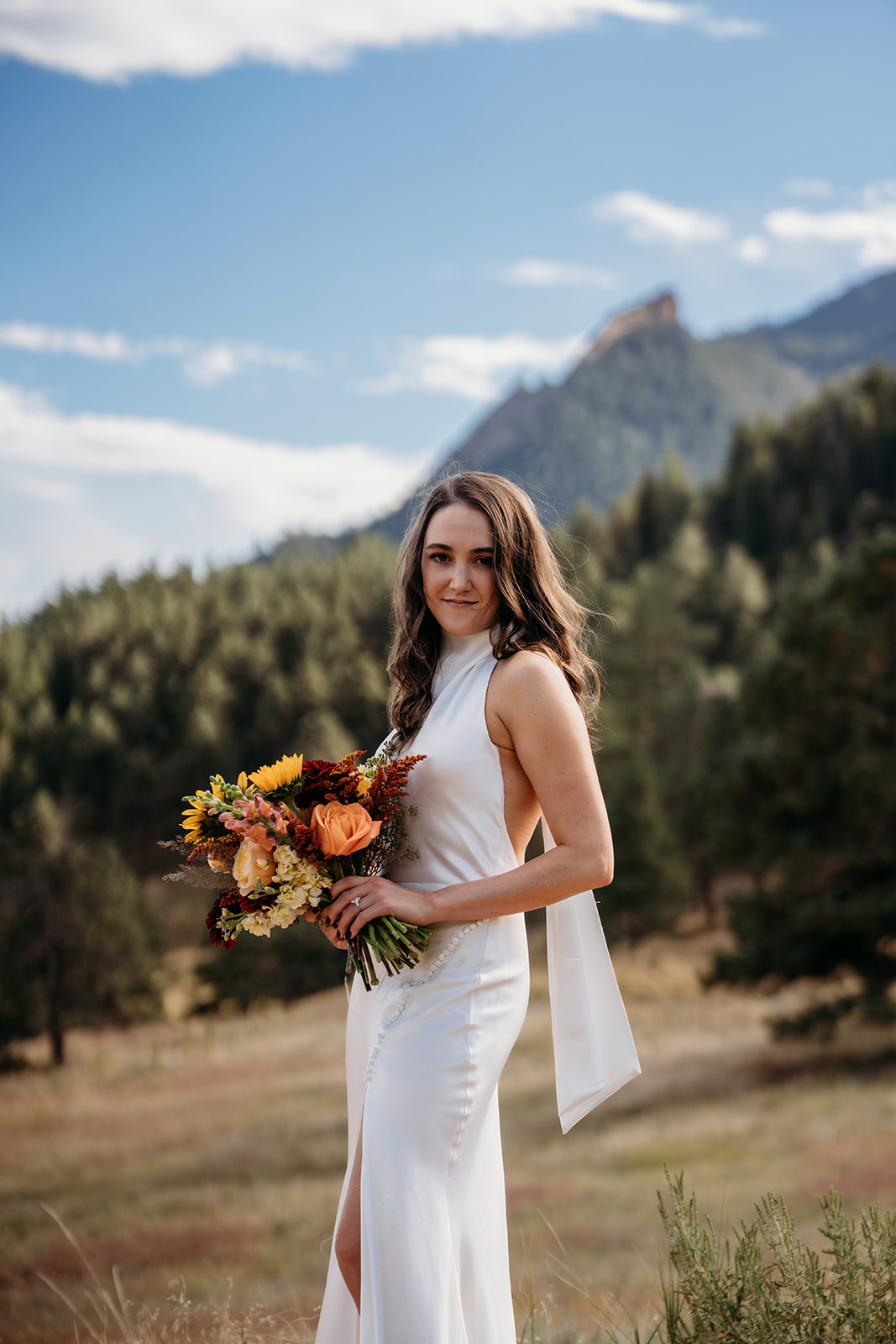 A bride holding a vibrant fall bouquet and smiling softly in a mountain meadow during her Colorado wedding day.