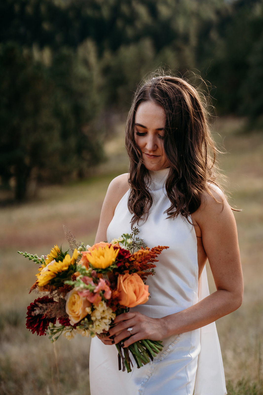 A bride holding a colorful fall bouquet and looking down thoughtfully during a quiet portrait moment in the Colorado foothills.