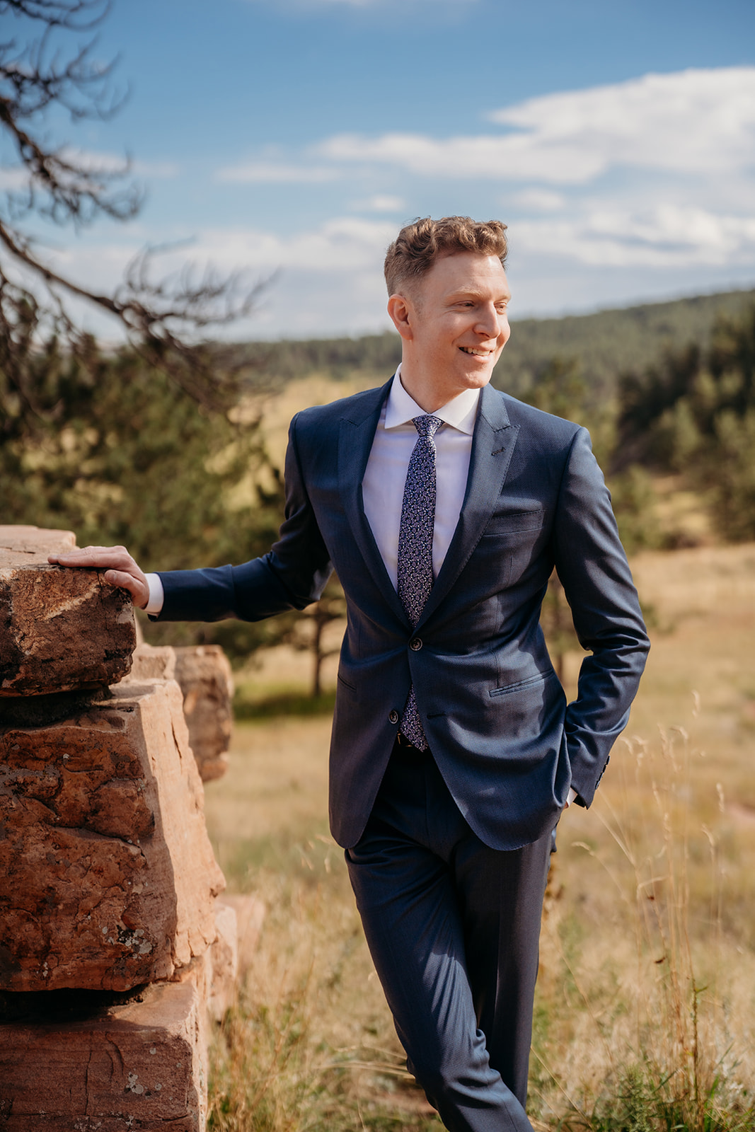 A groom leaning casually against a stone wall in the Colorado mountains, smiling during a relaxed wedding portrait.