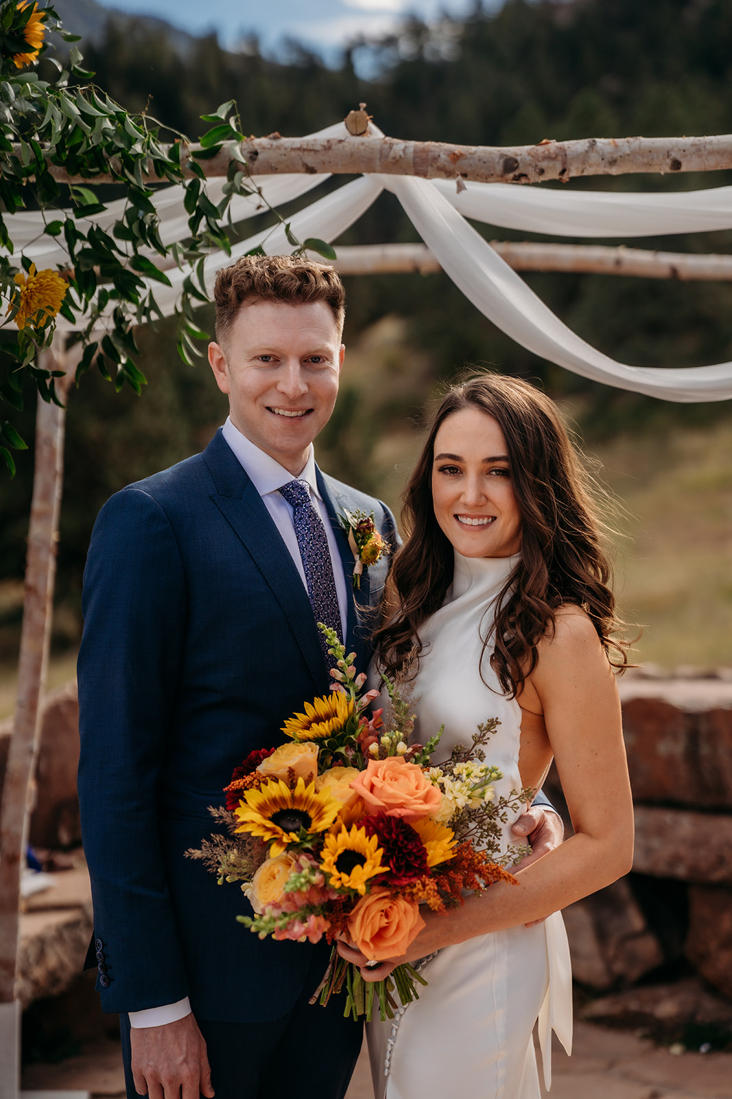 A bride and groom standing together beneath a floral ceremony arch, smiling toward the camera during an intimate Colorado wedding.
