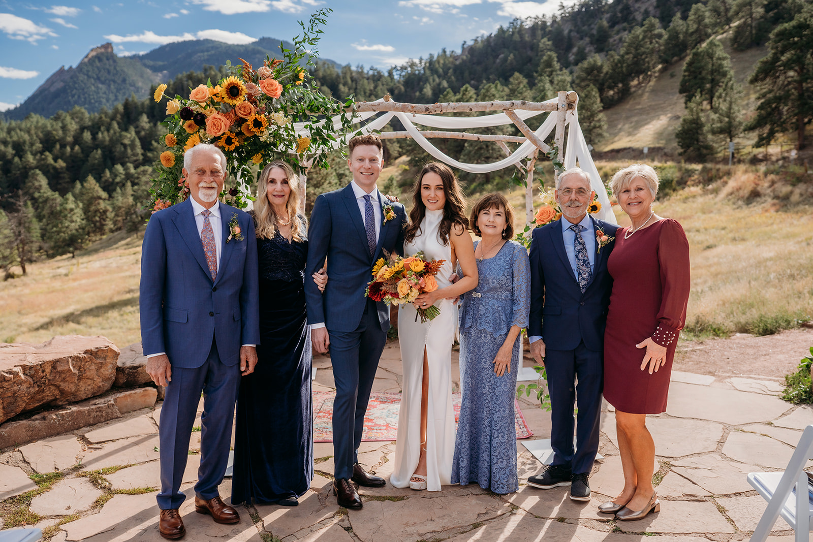 A large family portrait with the bride and groom standing beneath a floral arch in the Colorado foothills during their wedding day.