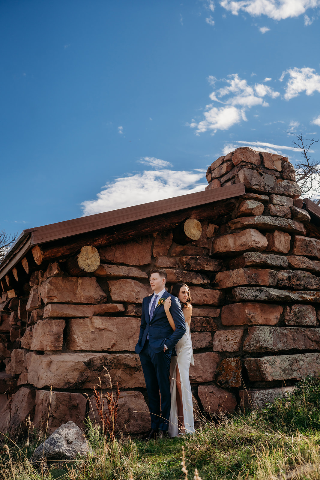 A bride and groom standing back to back against a rustic stone structure in the Colorado mountains on their wedding day.