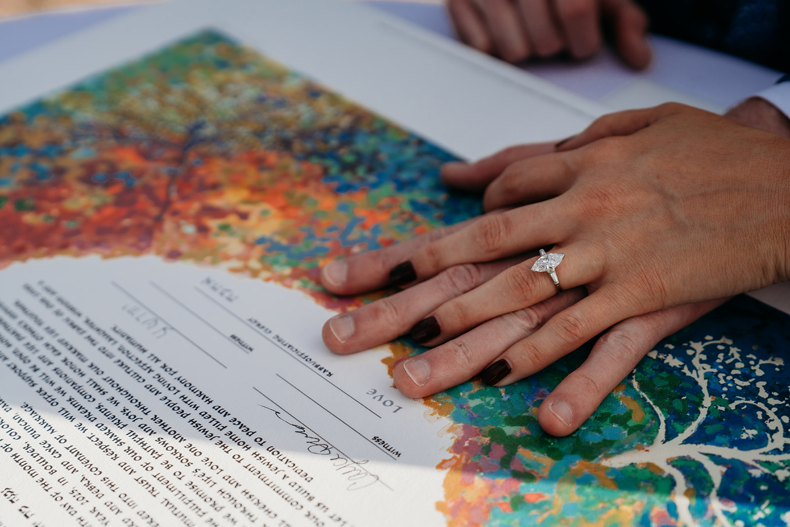 A close-up of hands resting on a colorful ketubah or wedding document, highlighting an engagement ring during an intimate Colorado wedding ceremony.