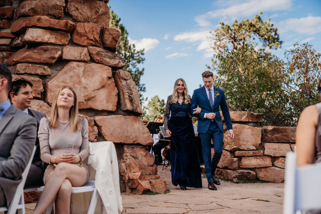 A couple walking together down a stone aisle between seated guests during an outdoor mountain wedding ceremony.