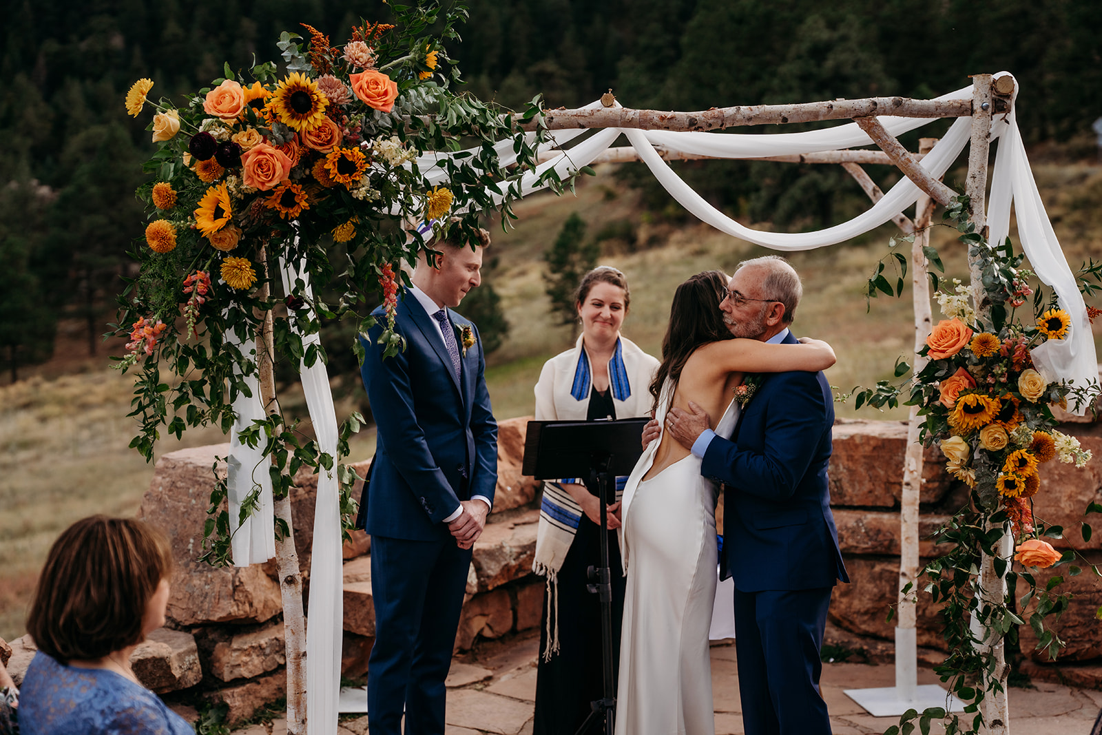 The bride hugging an older family member beneath the ceremony arch as the groom looks on during a heartfelt moment at a Colorado wedding.