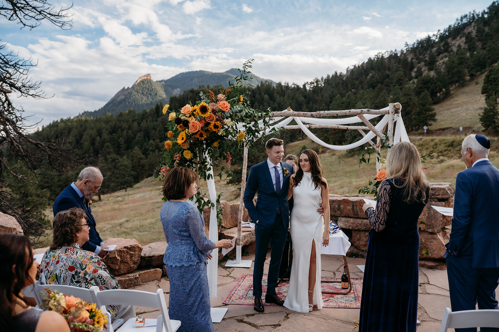 The newly married couple standing together under a floral arch as guests gather around them at an intimate mountain wedding ceremony.